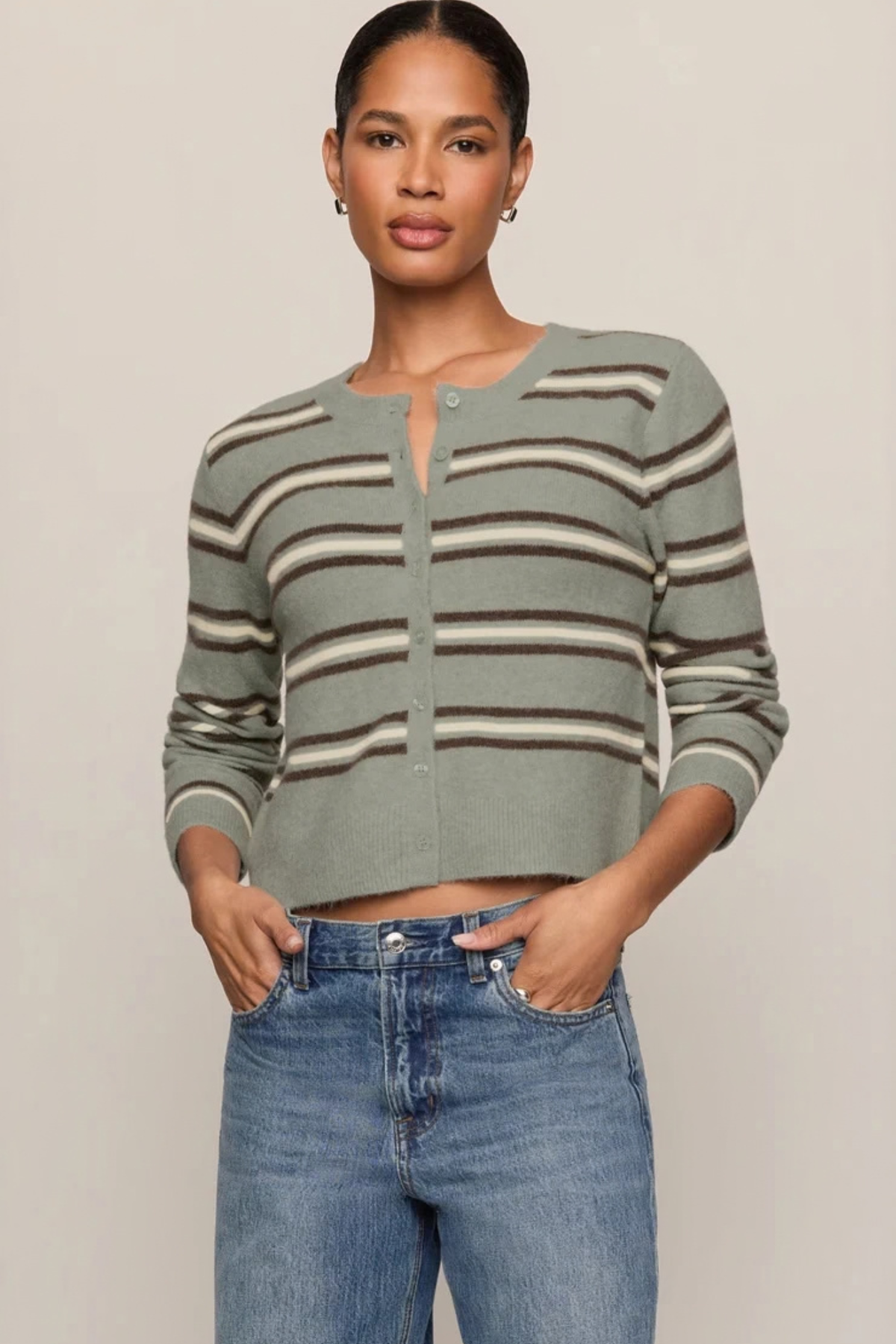 A woman modeling a cropped, long-sleeve knit cardigan with horizontal stripes in sage green, brown, and beige, paired with blue jeans against a plain beige studio background.