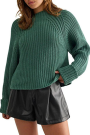 A woman wearing a chunky ribbed knit sweater in a medium green color, paired with black faux-leather shorts, shown against a plain white background.