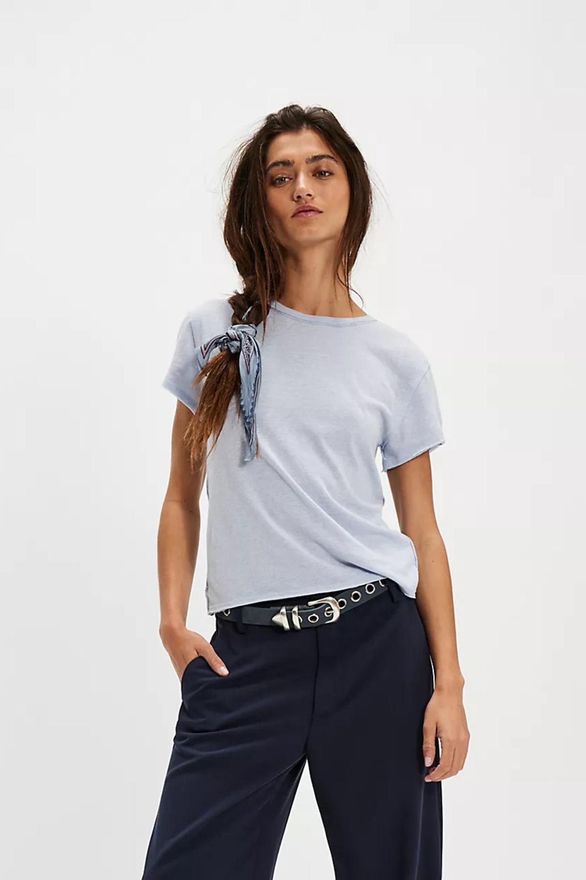 A woman modeling a light blue crew-neck t-shirt with a blue patterned scarf tied to the left shoulder, paired with dark navy trousers and a black belt with silver studs, against a plain white studio background.