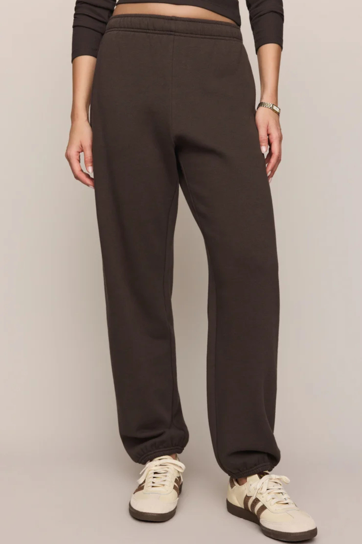 A person wearing dark brown sweatpants with a drawstring waist and elastic cuffs, paired with cream sneakers, shown from the waist down against a plain light gray studio background.