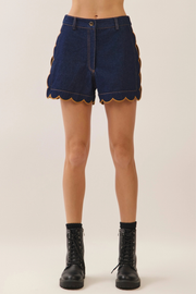 A person wearing dark blue denim shorts with scalloped edges and gold trim, paired with black ankle boots, shown against a plain light beige studio background.