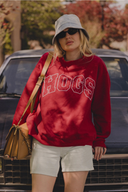 A woman wearing a red sweatshirt with 'HOGS' in white varsity letters, paired with a gray bucket hat, sunglasses, white shorts, and a tan shoulder bag, standing in front of a dark car outdoors.