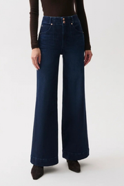 A model wearing dark blue wide-leg denim jeans with a high waist, paired with a dark brown long-sleeve top and dark brown pointed-toe boots, against a plain white studio background.