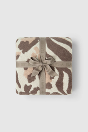 A folded plush throw blanket with a zebra-like animal print pattern in brown, beige, and cream, wrapped with a taupe ribbon that has the BARFOOT BAR brand name printed on it, set against a light gray background.