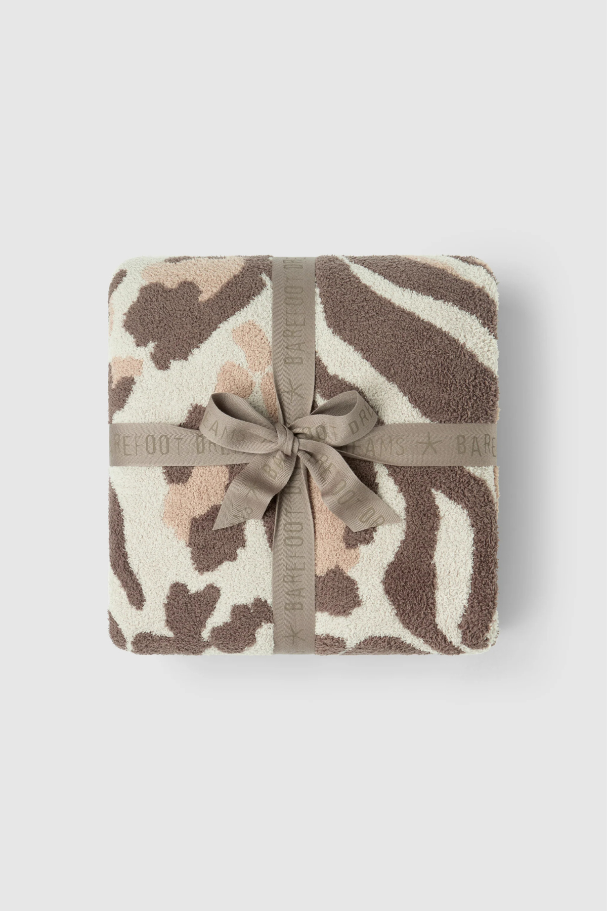 A folded plush throw blanket with a zebra-like animal print pattern in brown, beige, and cream, wrapped with a taupe ribbon that has the BARFOOT BAR brand name printed on it, set against a light gray background.