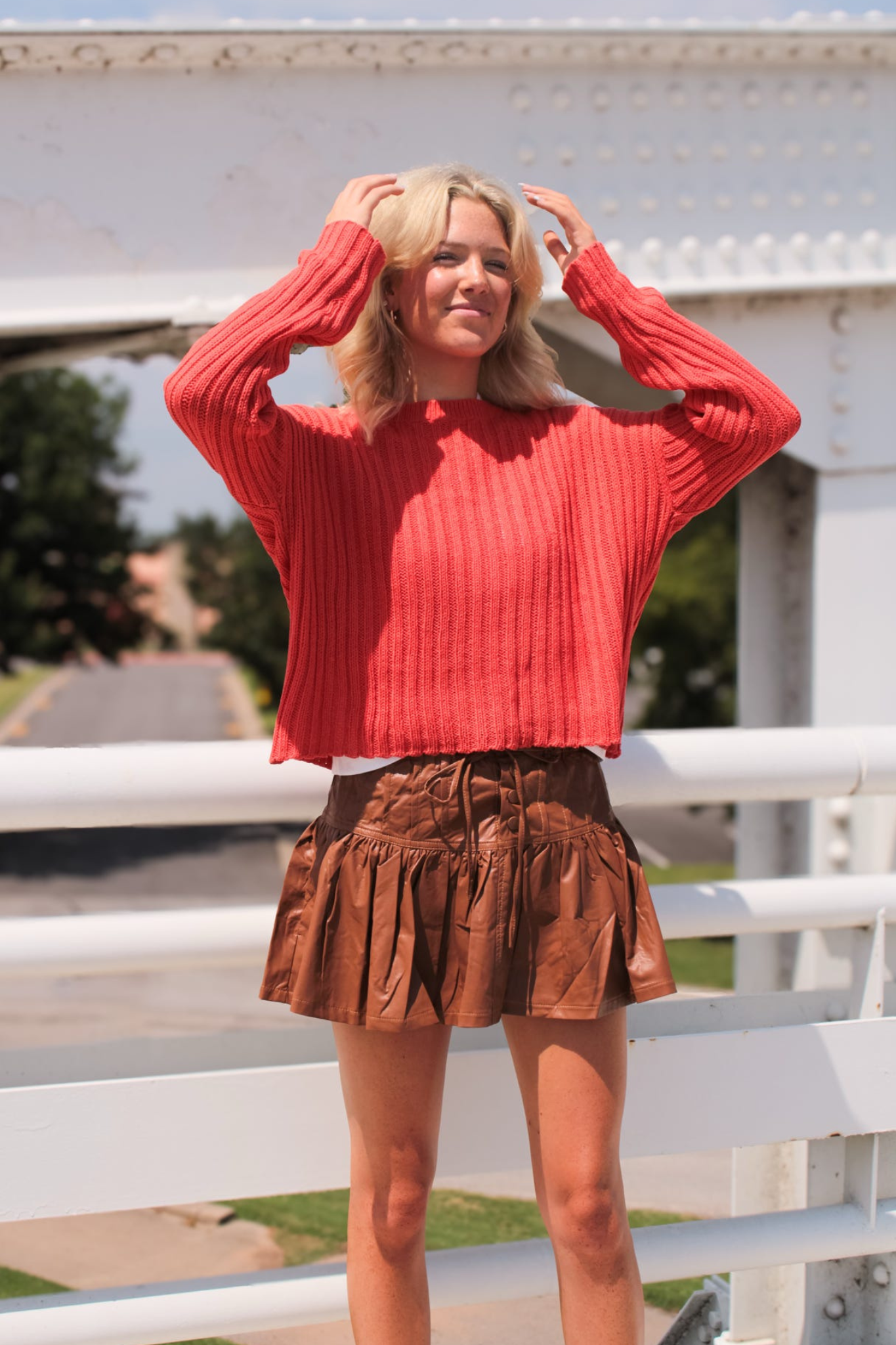 Woman wearing a red sweater and brown skirt standing in front of a white fence.