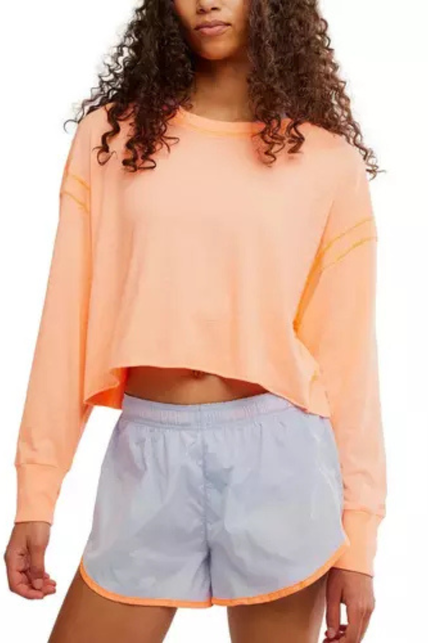 A model wearing a cropped, long-sleeve peach-orange sweatshirt and light blue athletic shorts with a peach-orange trim, shown against a plain white background.