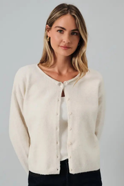 A woman wearing a cream-colored, soft knit cardigan with a button-up front over a white ribbed top and black pants, photographed against a solid light gray background.