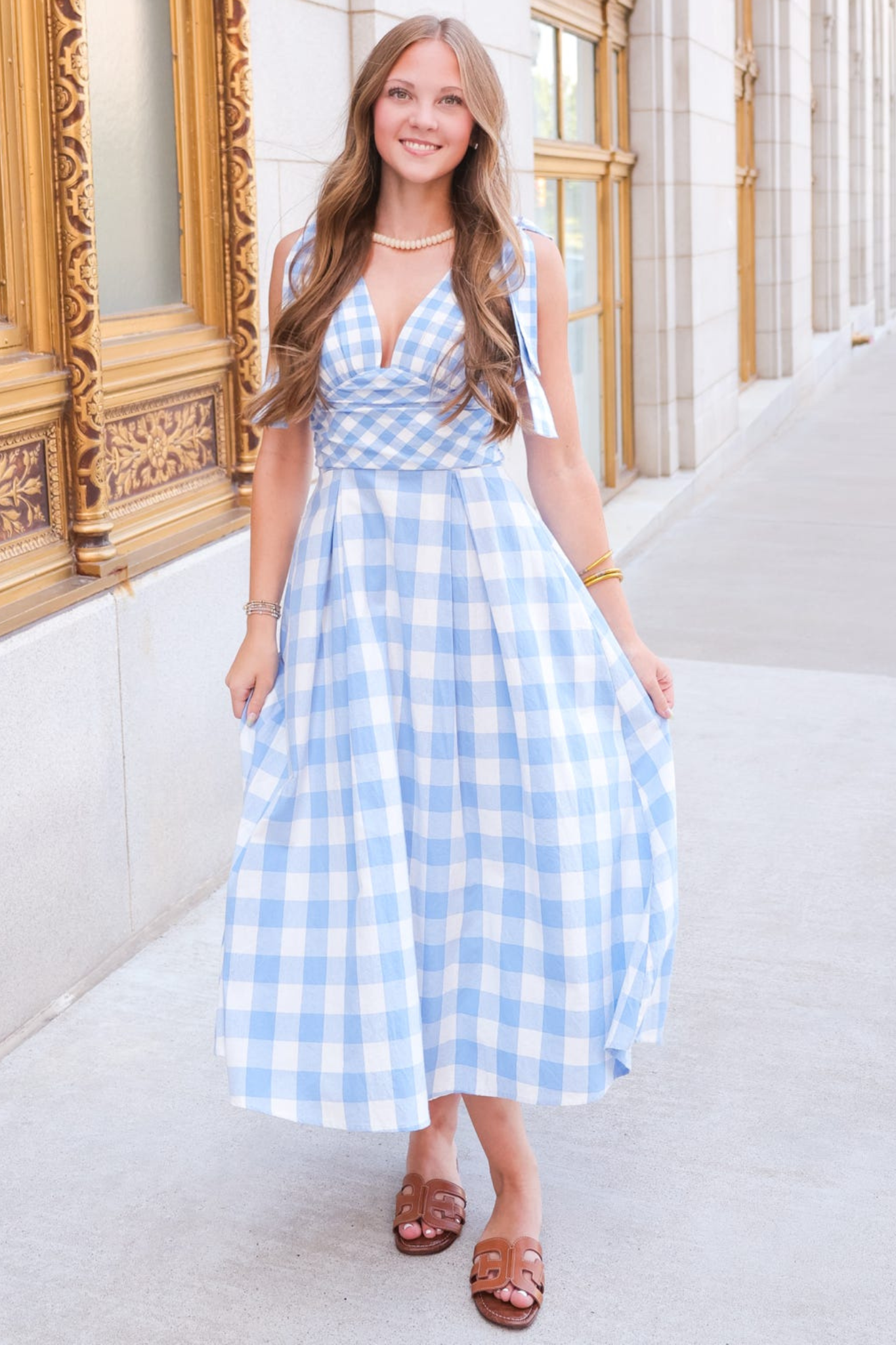 A woman wearing a light blue and white gingham midi dress with a tie-shoulder design, standing on a sidewalk in front of a building with ornate gold-framed windows.
