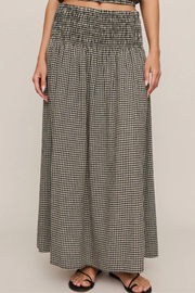 A woman wearing a long black and white gingham maxi skirt with a smocked waistband, paired with black strappy sandals, shown against a solid light beige background.
