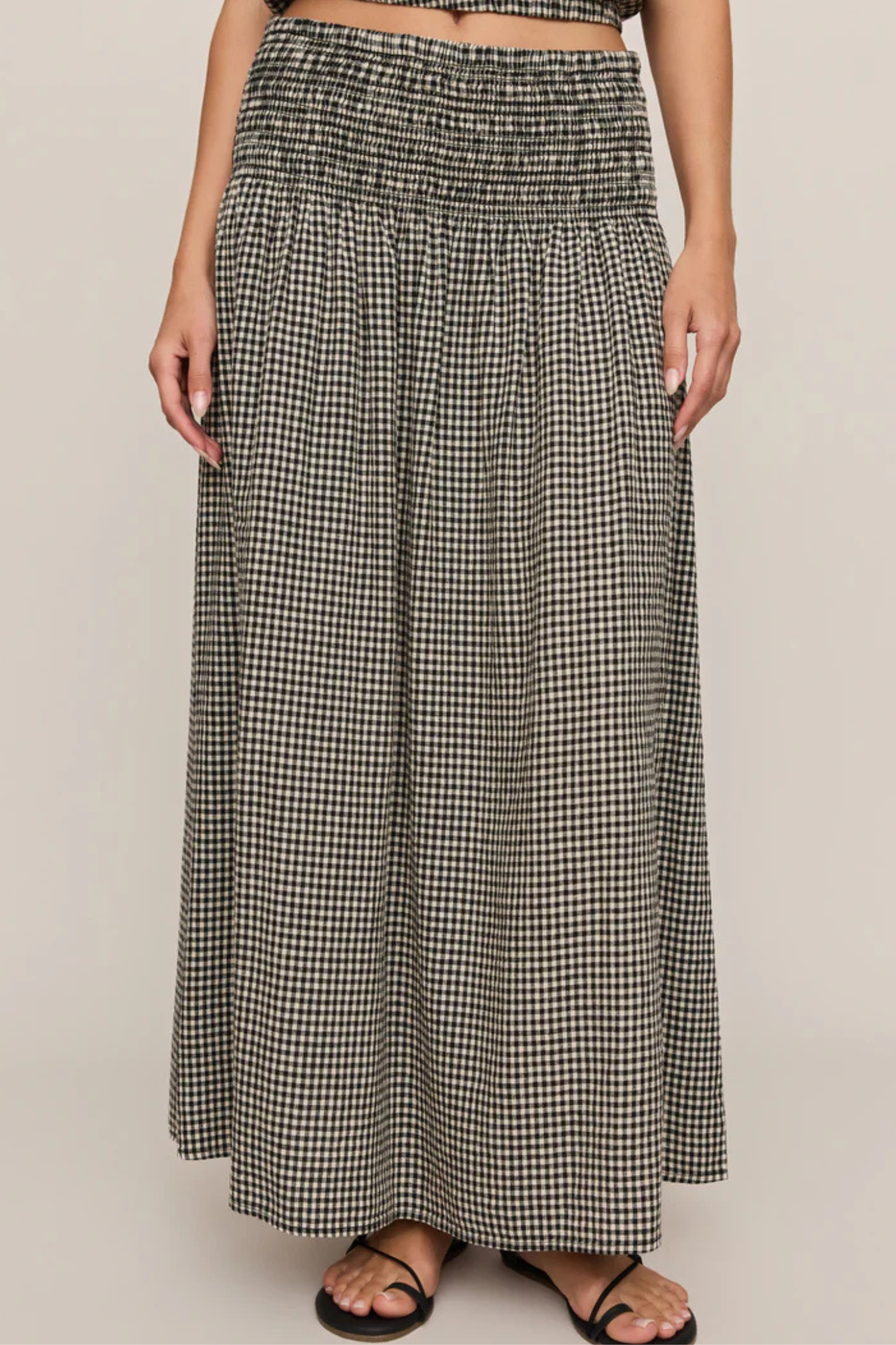 A woman wearing a long black and white gingham maxi skirt with a smocked waistband, paired with black strappy sandals, shown against a solid light beige background.