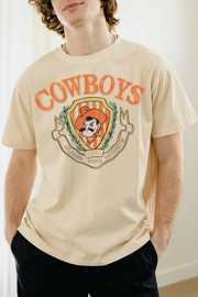 A man wearing a cream-colored t-shirt with a vintage-style orange and brown 'COWBOYS' graphic on the front, paired with black pants.