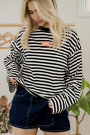A woman wearing a black and white horizontally striped long-sleeve sweatshirt with a small orange rectangular chest patch, paired with dark denim shorts.