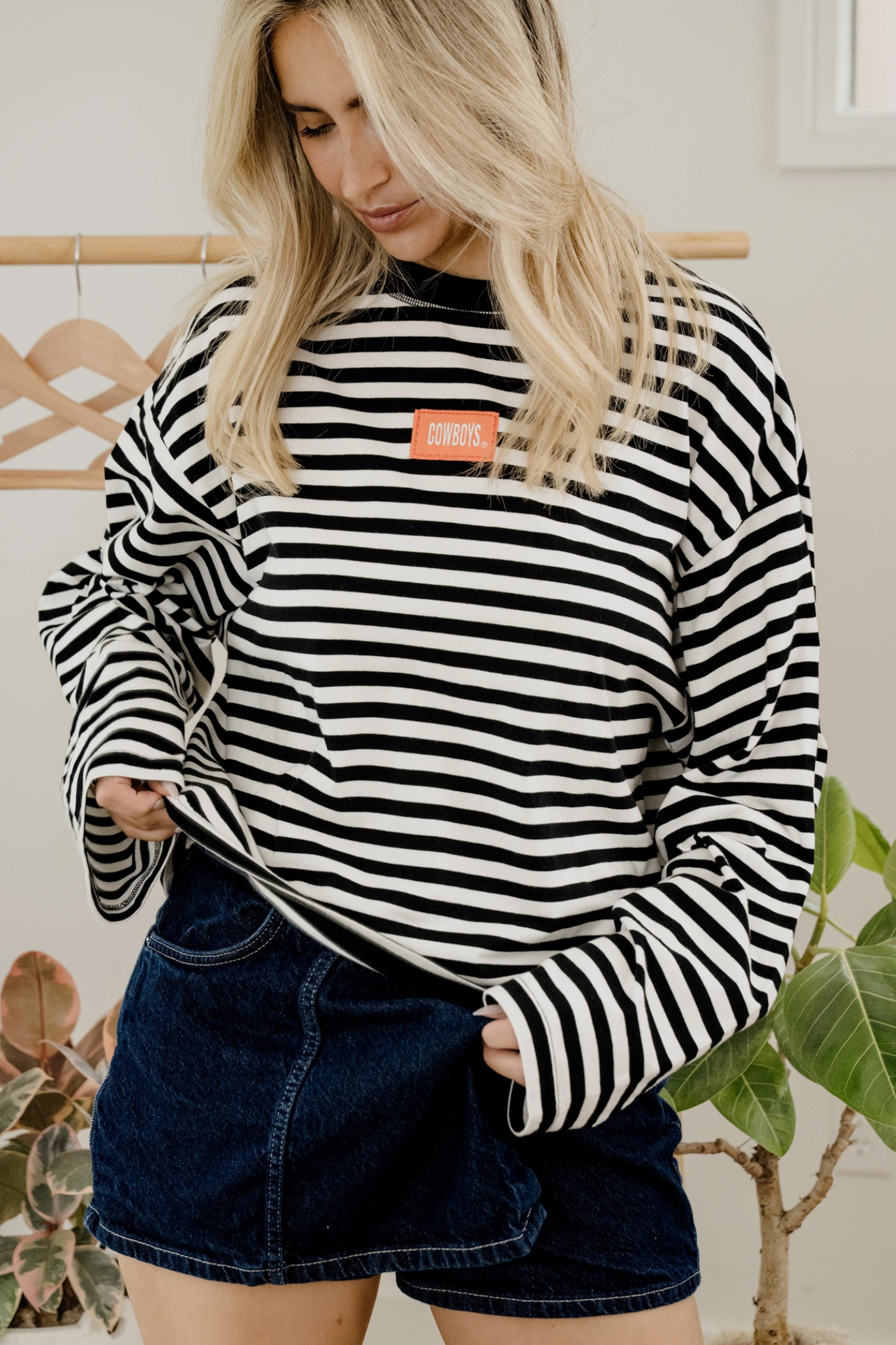 A woman wearing a black and white horizontally striped long-sleeve sweatshirt with a small orange rectangular chest patch, paired with dark denim shorts.