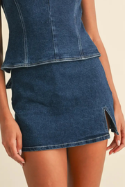 A close-up of a model wearing a dark blue denim mini skirt with a side slit and a matching cropped denim top against a light beige background.