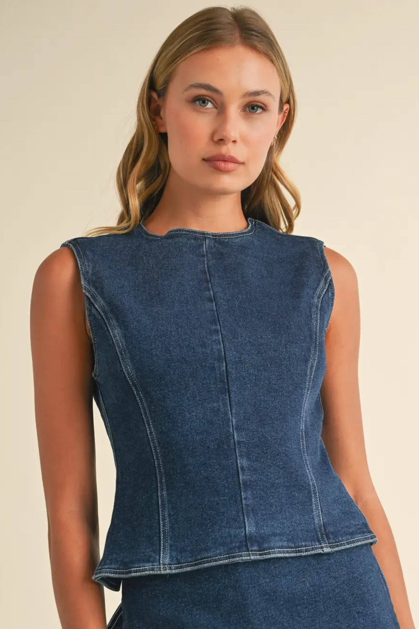 A woman wearing a sleeveless dark blue denim top with a structured, corset-style bodice and a slightly flared hem, photographed against a plain beige studio background.