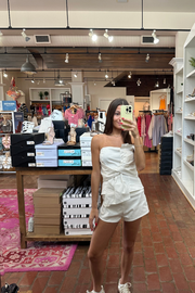 A woman takes a mirror selfie in a clothing boutique, wearing a white strapless ruffled romper and white sneakers. The store features brick floors, wooden ceiling beams, and racks of clothing in the background.