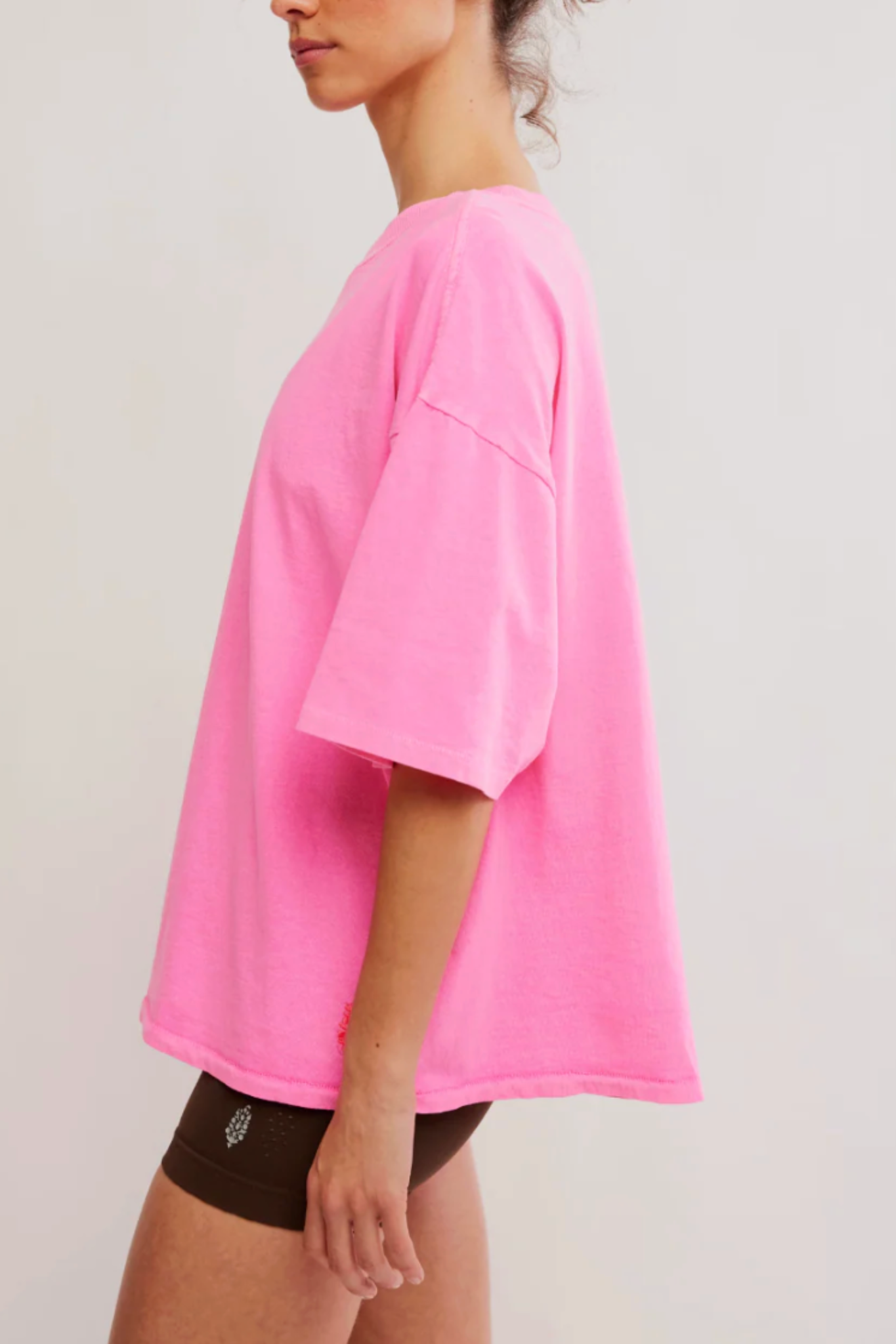 A woman shown from the side wearing an oversized bright pink short-sleeve T-shirt and dark brown shorts against a plain light gray studio background.