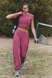 A woman walks on a grassy tennis court wearing a matching dusty rose athletic set consisting of a short-sleeve crop top and wide-leg sweatpants, holding a tennis racket.