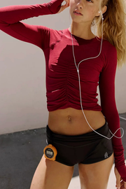A woman wearing a fitted long-sleeve ruched athletic top in deep burgundy red and black running shorts, with white earphones and a small orange stopwatch clipped to her waist.