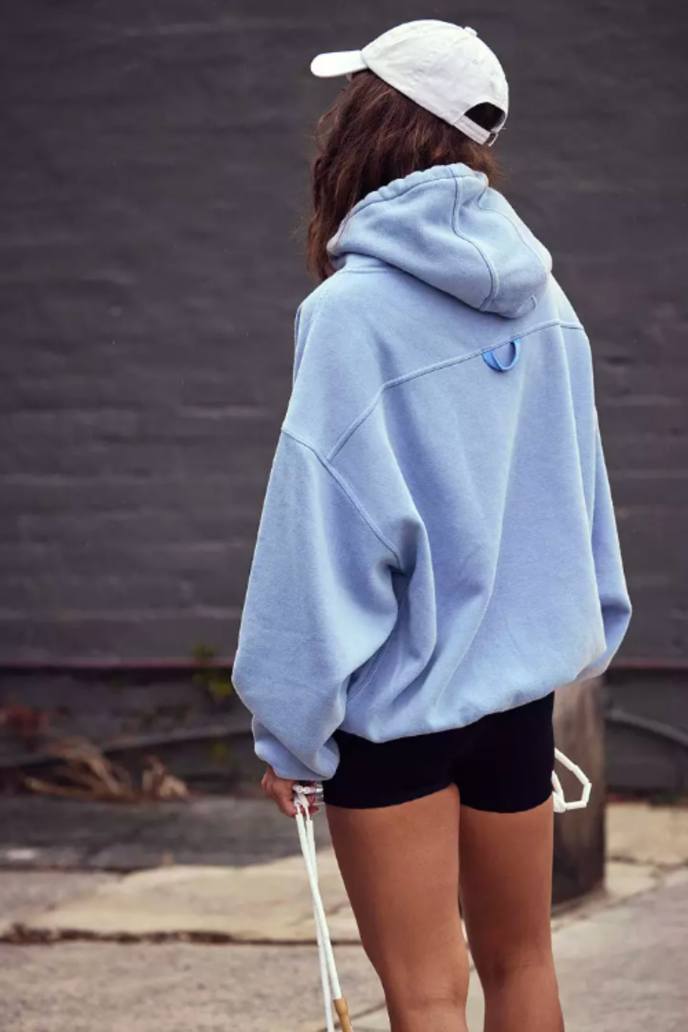 A person shown from the back wearing an oversized light blue hoodie, black shorts, and a white baseball cap, standing outdoors against a dark gray wall.