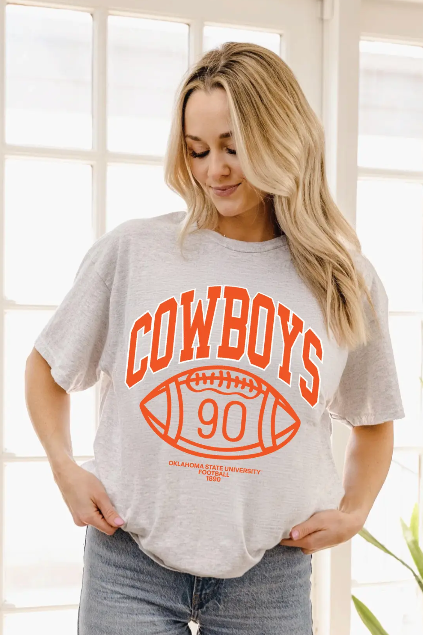 A woman wearing a light heather gray t-shirt with a bright orange graphic that reads 'COWBOYS' above a football-shaped '90' and 'OKLAHOMA STATE UNIVERSITY FOOTBALL'.