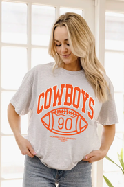 A woman wearing a light heather gray t-shirt with a bright orange graphic that reads 'COWBOYS' above a football-shaped '90' and 'OKLAHOMA STATE UNIVERSITY FOOTBALL'.