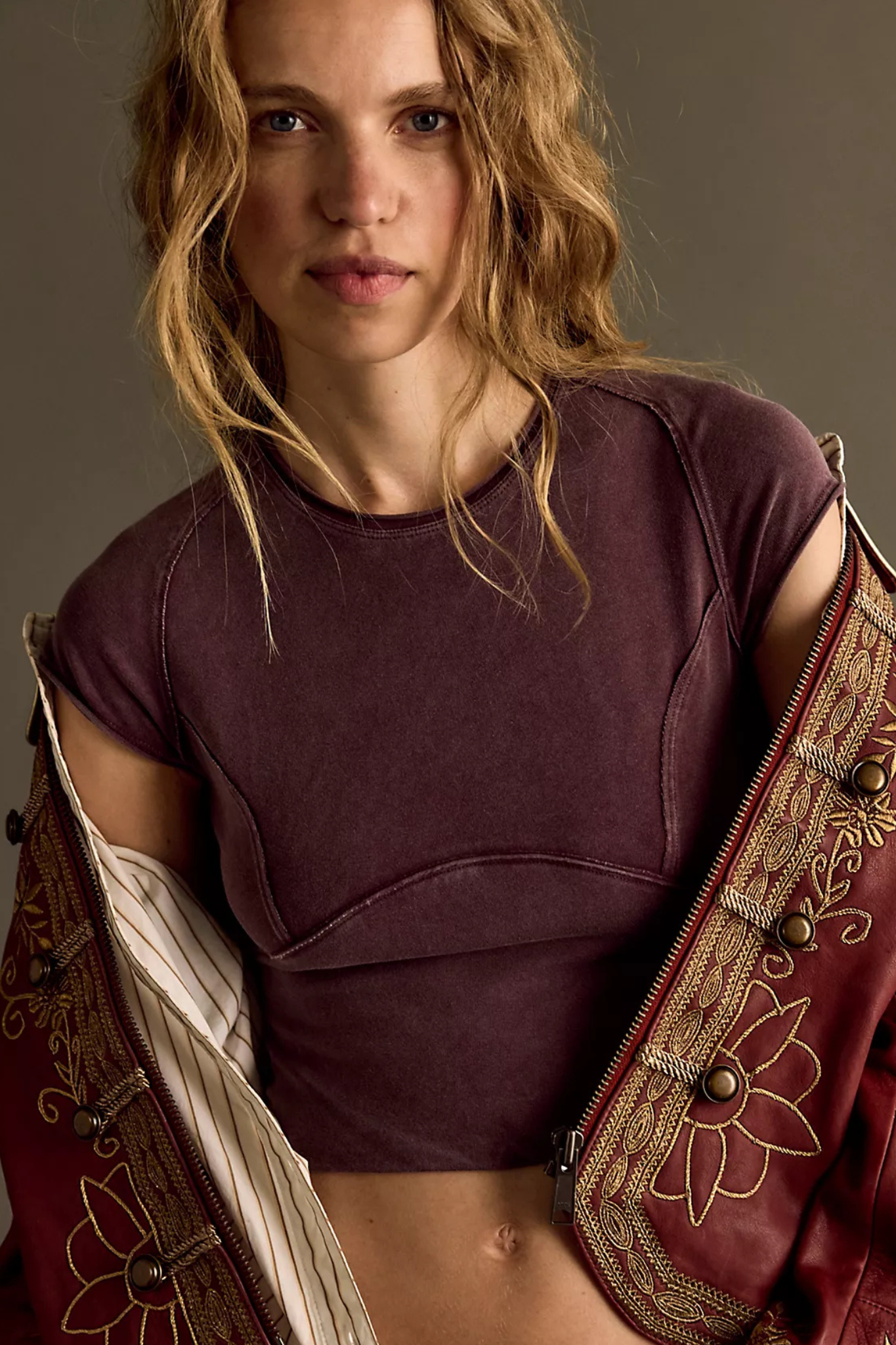 A woman wearing a dark plum purple cropped t-shirt with unique seam detailing, styled with a red embroidered jacket draped over her shoulders, set against a dark brown studio background.