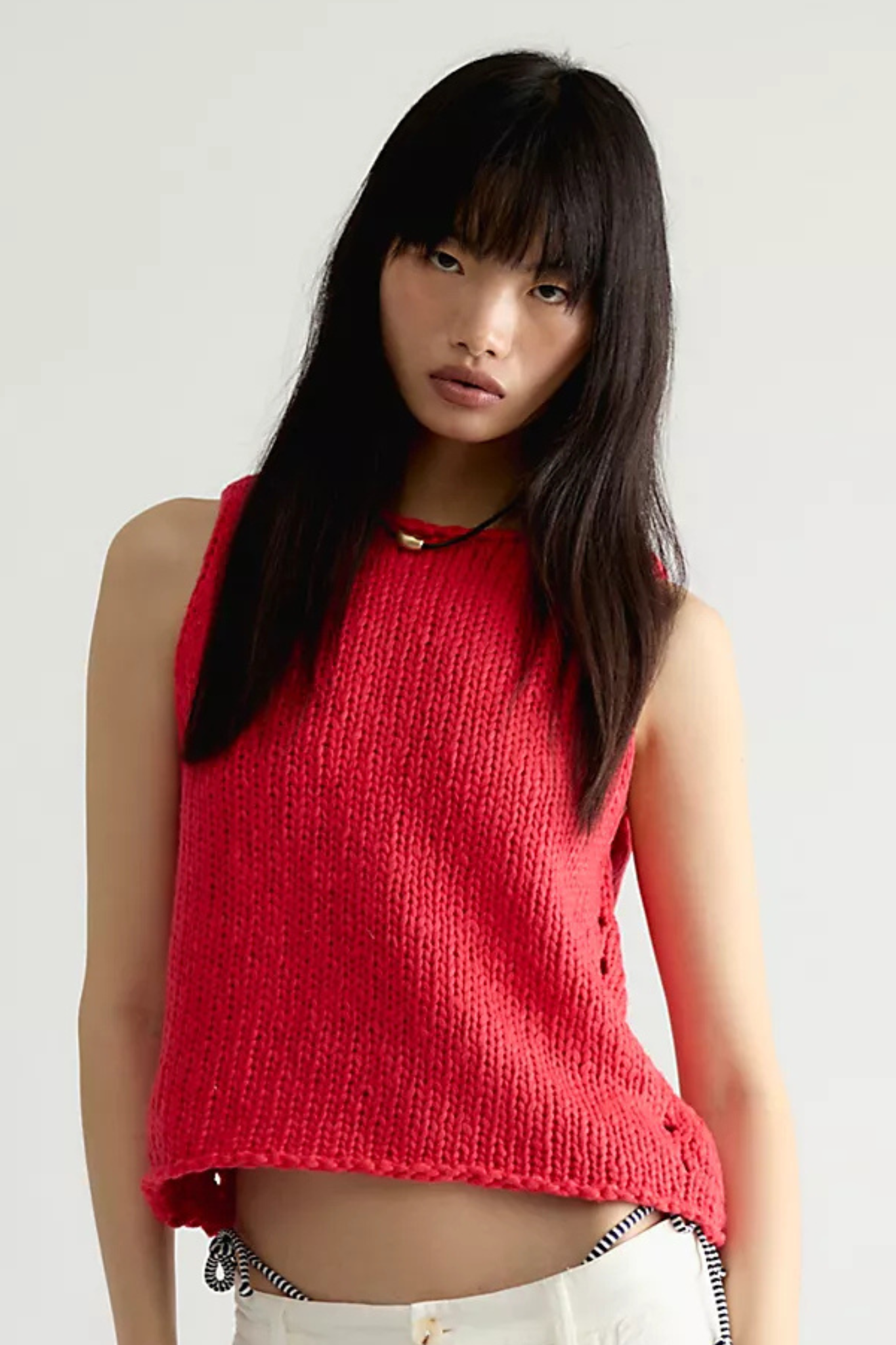 A model wearing a vibrant red sleeveless knit top with a crew neck and side buttons, paired with white pants, against a plain light gray background.
