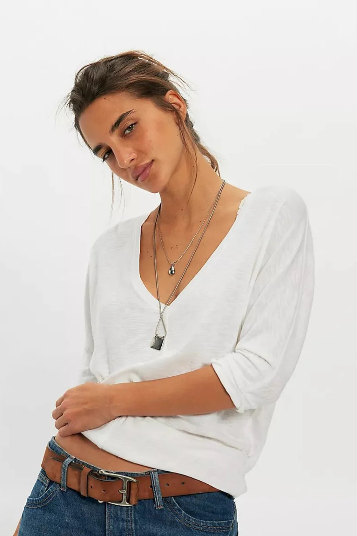 A woman wearing a white long-sleeve V-neck top tucked into blue jeans with a brown belt, accessorized with layered silver necklaces, against a plain white studio background.