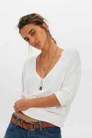 A woman wearing a white long-sleeve V-neck top tucked into blue jeans with a brown belt, accessorized with layered silver necklaces, against a plain white studio background.