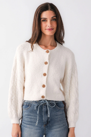 A woman wearing a cream-colored, textured fuzzy cardigan with brown buttons, paired with blue jeans, against a plain white studio background.