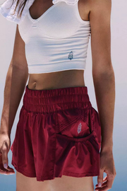 A close-up of a model wearing a pair of shiny burgundy athletic shorts with a gathered elastic waist and side pocket, paired with a white ribbed crop top.