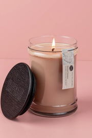A lit soy wax candle in a clear glass jar with a dark round wooden lid leaning beside it on a soft pink background. The tag reads 'BROKEN WAVE CANDLE CO.