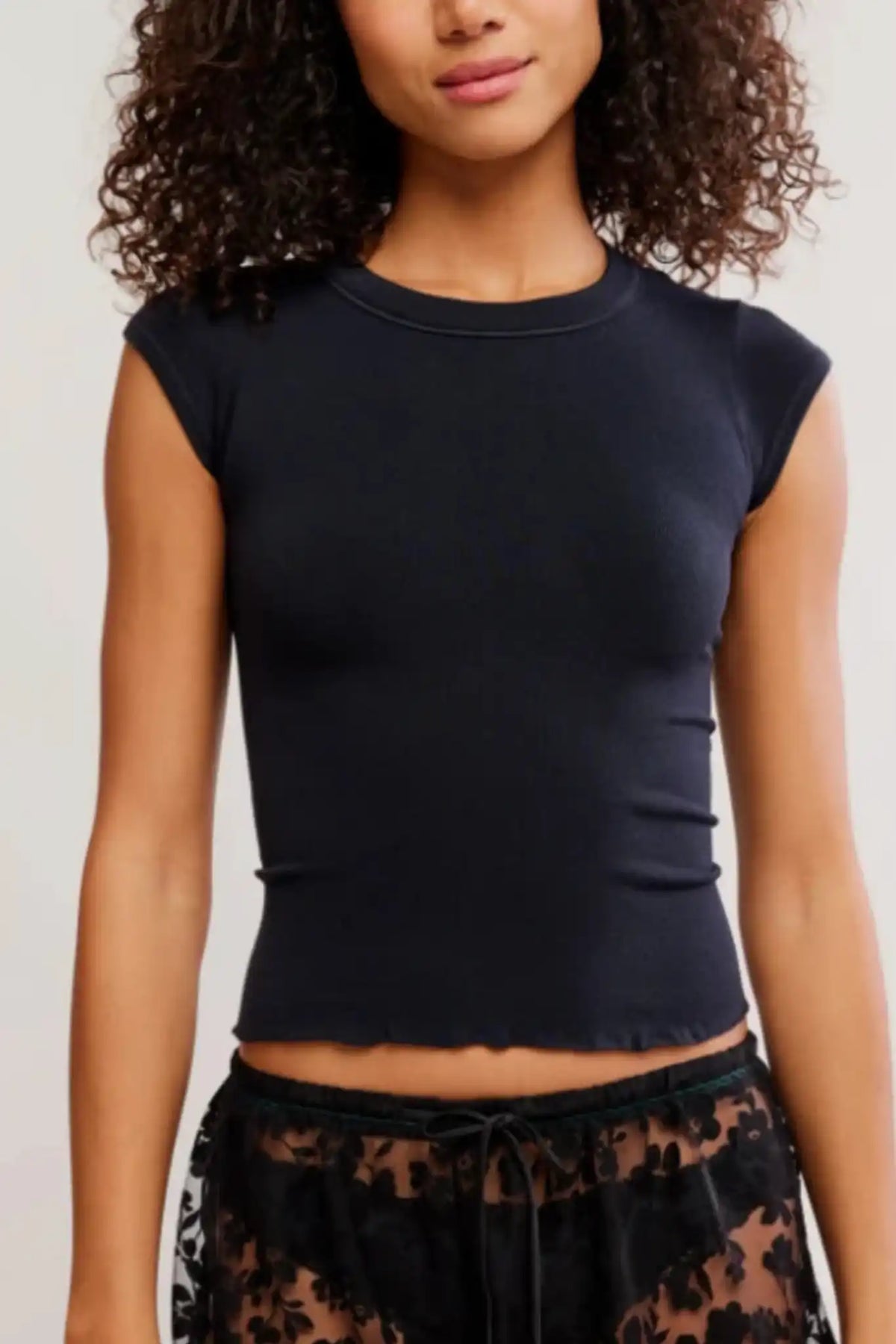 A woman wearing a fitted black short-sleeve crop top with a crew neckline, paired with black lace-patterned bottoms, against a plain light background.