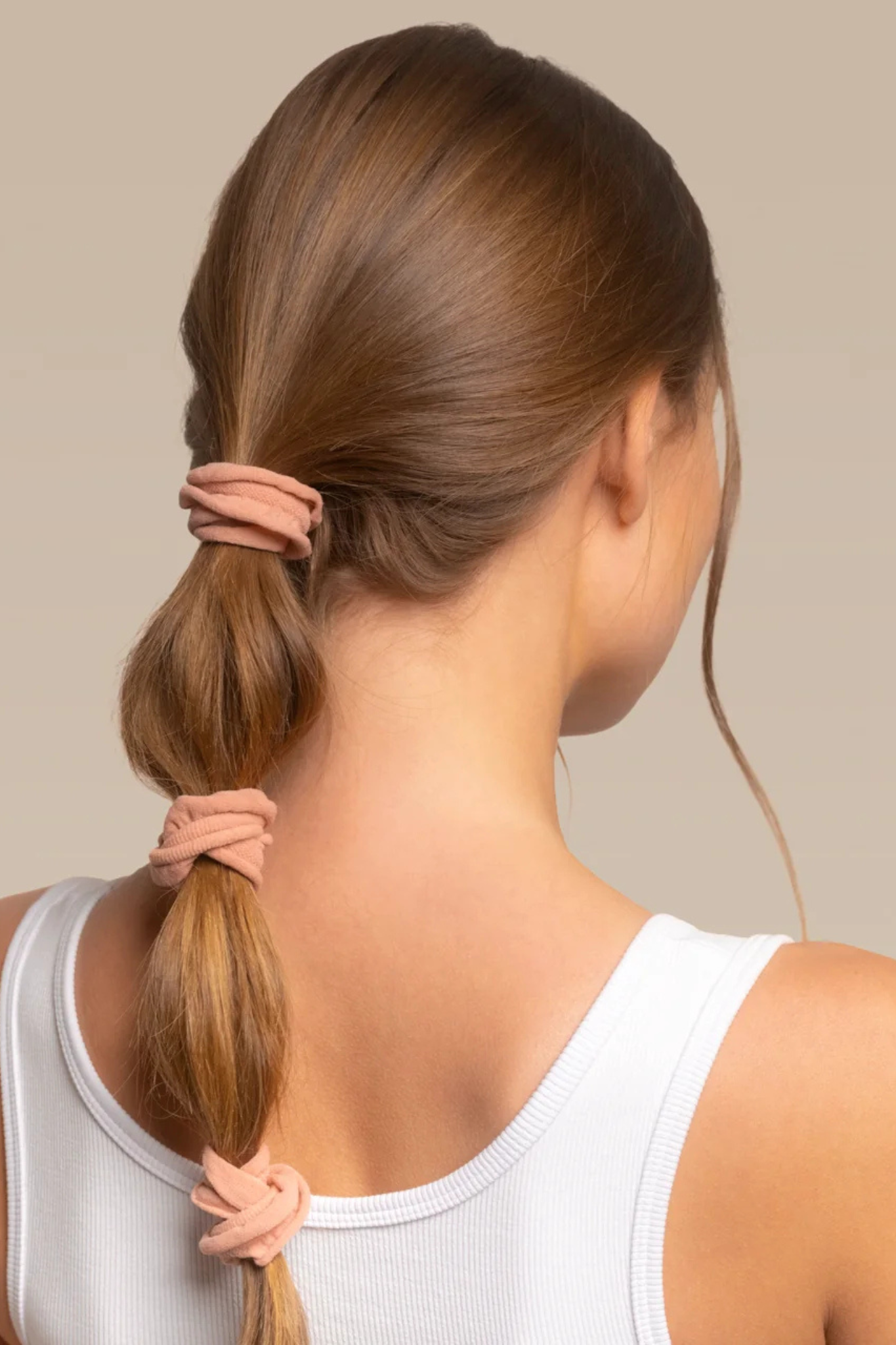 A woman shown from the back wearing a white ribbed tank top, demonstrating three dusty pink fabric scrunchie hair ties on her brown hair against a solid beige background.