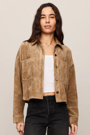 A woman modeling a cropped tan suede jacket over a white tank top and black jeans, photographed against a plain light beige studio background.