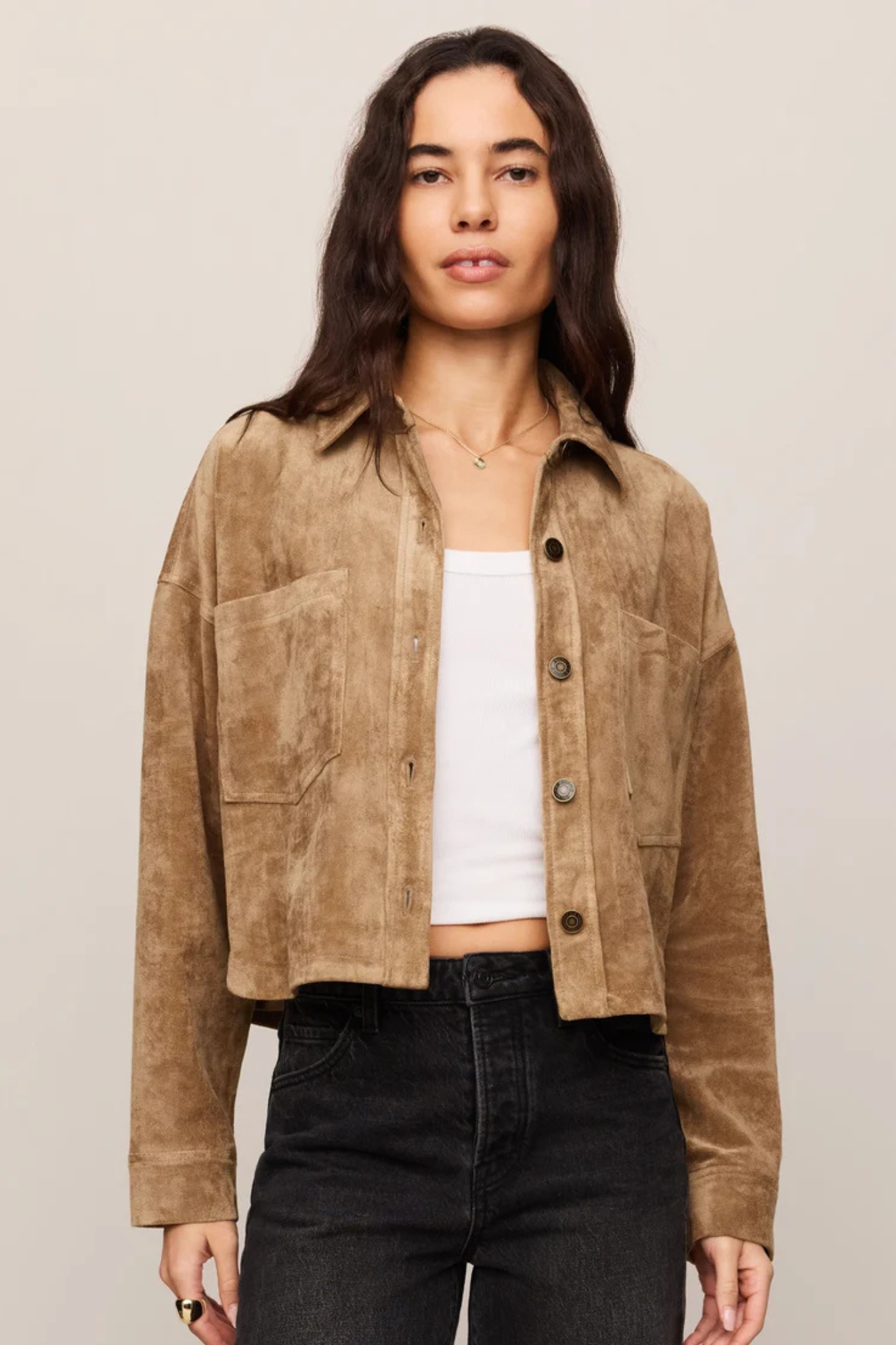A woman modeling a cropped tan suede jacket over a white tank top and black jeans, photographed against a plain light beige studio background.