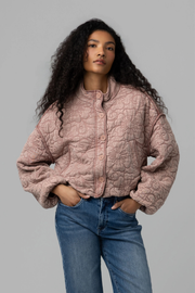 A model with long, curly black hair is wearing a dusty rose pink quilted jacket with a button-up front, paired with blue jeans, against a solid gray studio background.