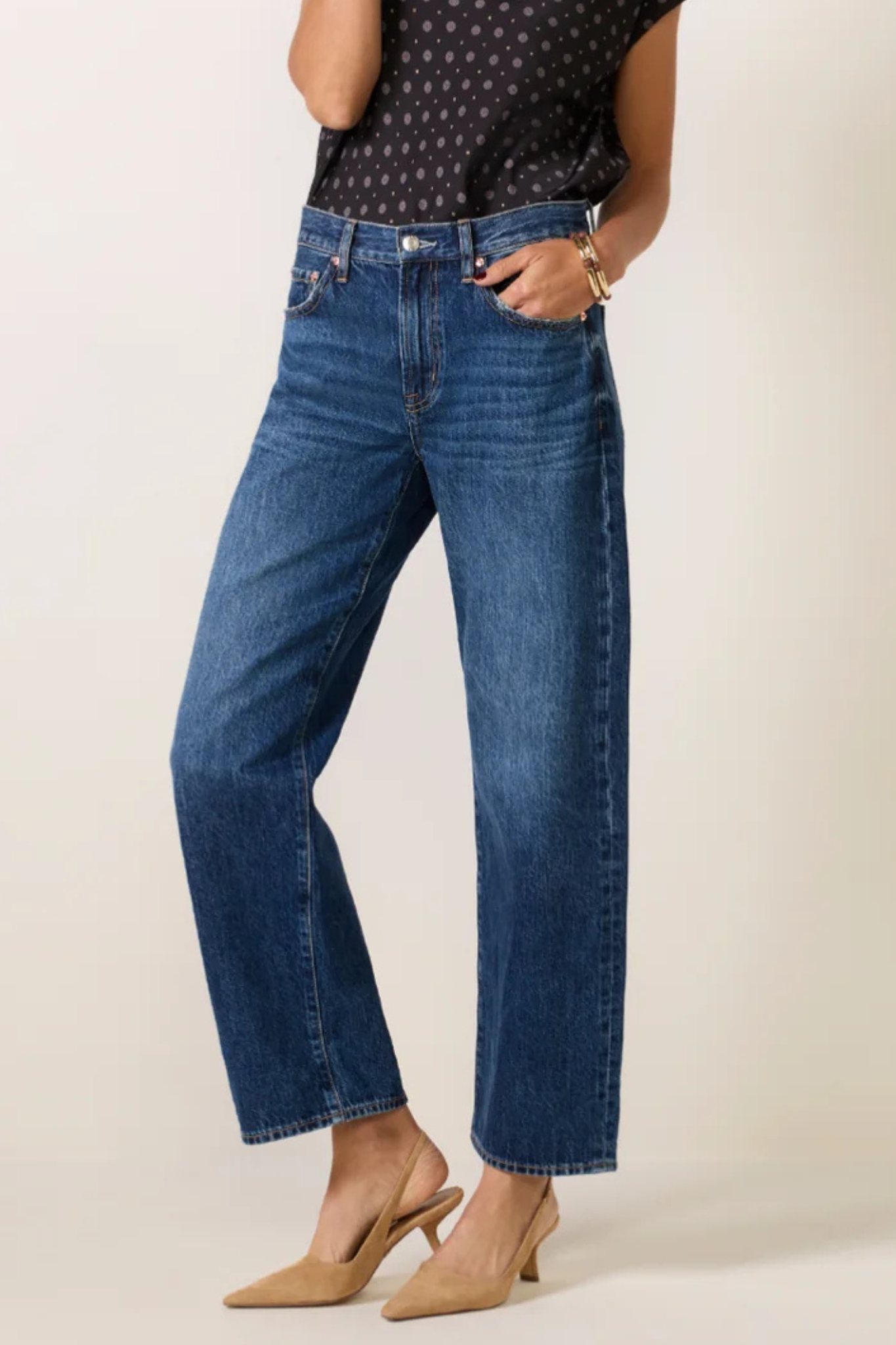 A model wearing a pair of medium-wash blue denim jeans with a straight-leg or wide-leg fit, paired with a black patterned top and beige slingback heels, against a plain light beige studio background.