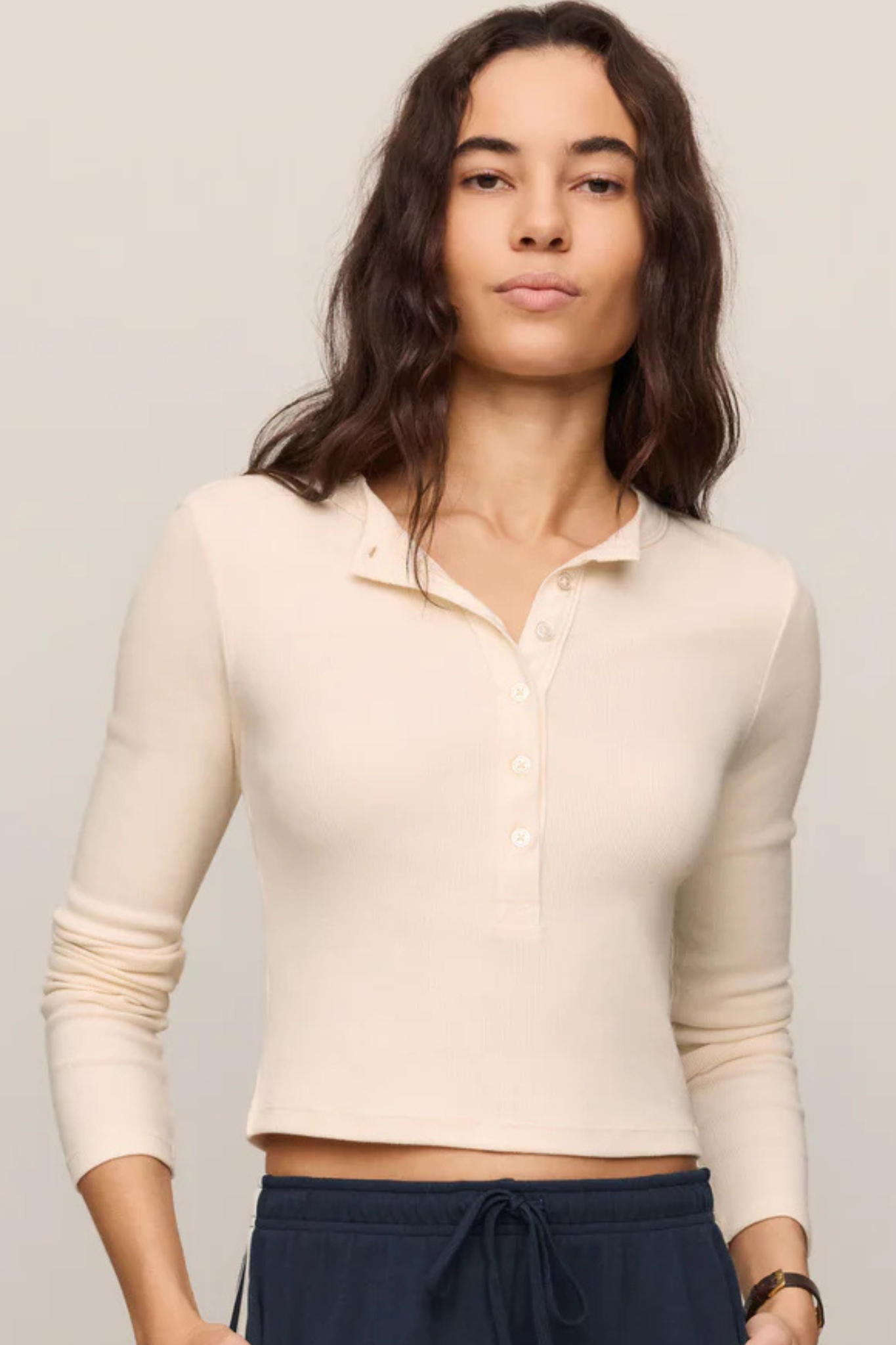 A woman wearing a cream-colored, long-sleeve ribbed knit polo henley top paired with navy blue drawstring pants, photographed against a solid light gray background.
