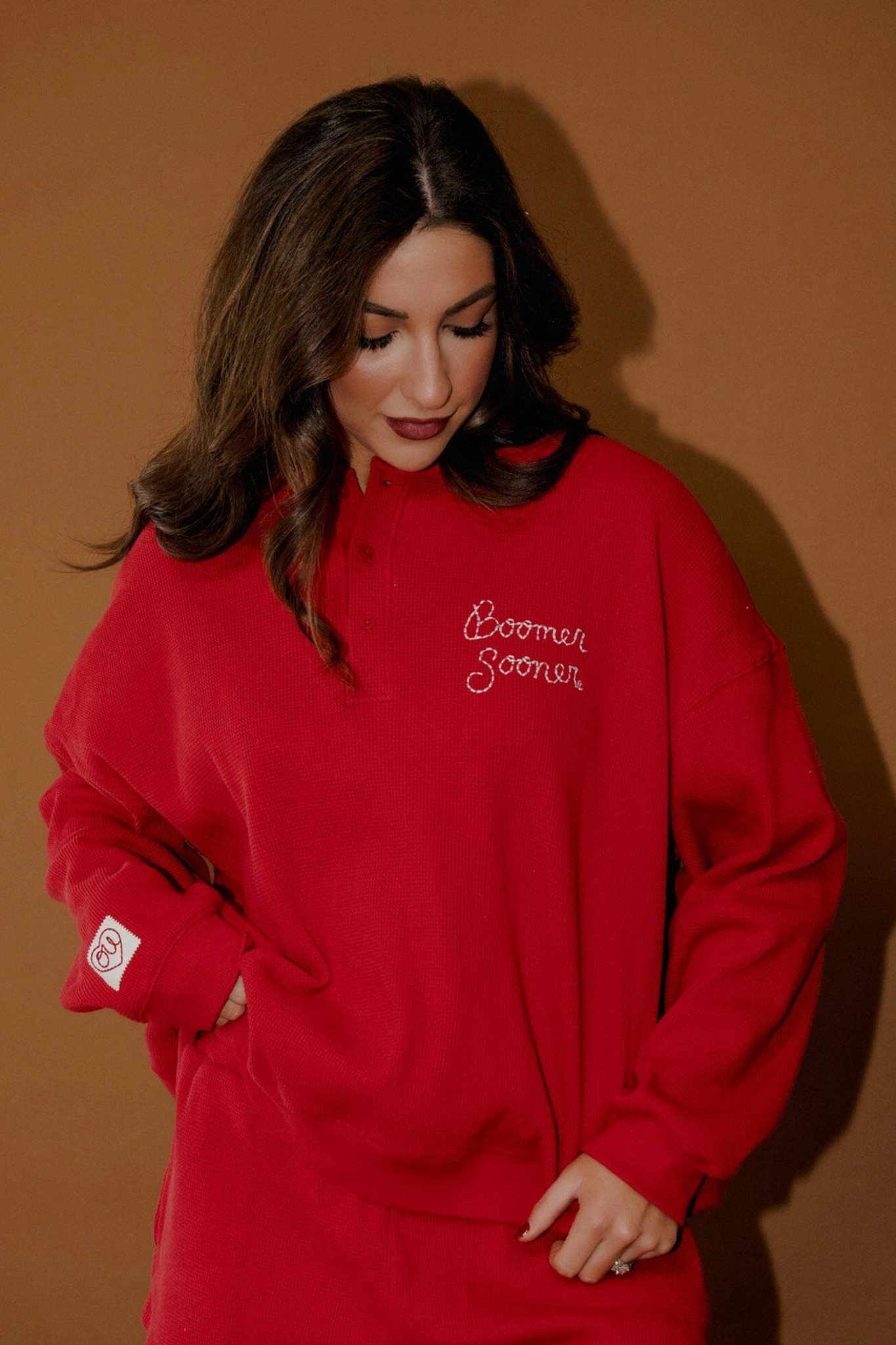 A woman wearing a bright red sweatshirt with 'Boomer Later' embroidered in white script on the left chest, posed against a solid warm brown background.