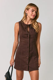 A woman modeling a sleeveless brown corduroy mini dress with a button-down front and a tie-neck detail, set against a plain light gray background.