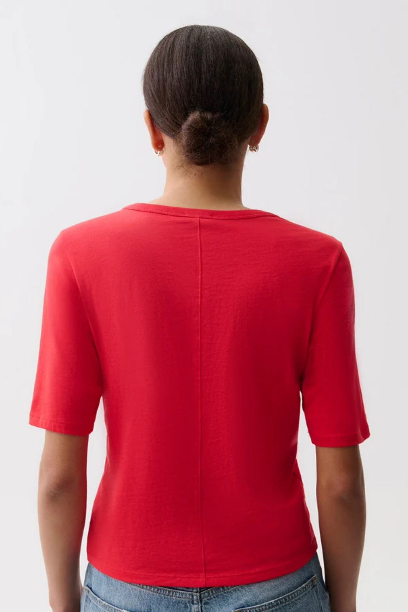 A woman viewed from the back wearing a bright red short-sleeve crewneck top and light blue jeans against a plain light gray studio background.