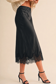 A black satin midi skirt with a lace hem, shown on a model wearing a cream knit sweater and black high-heeled sandals against a warm beige background.
