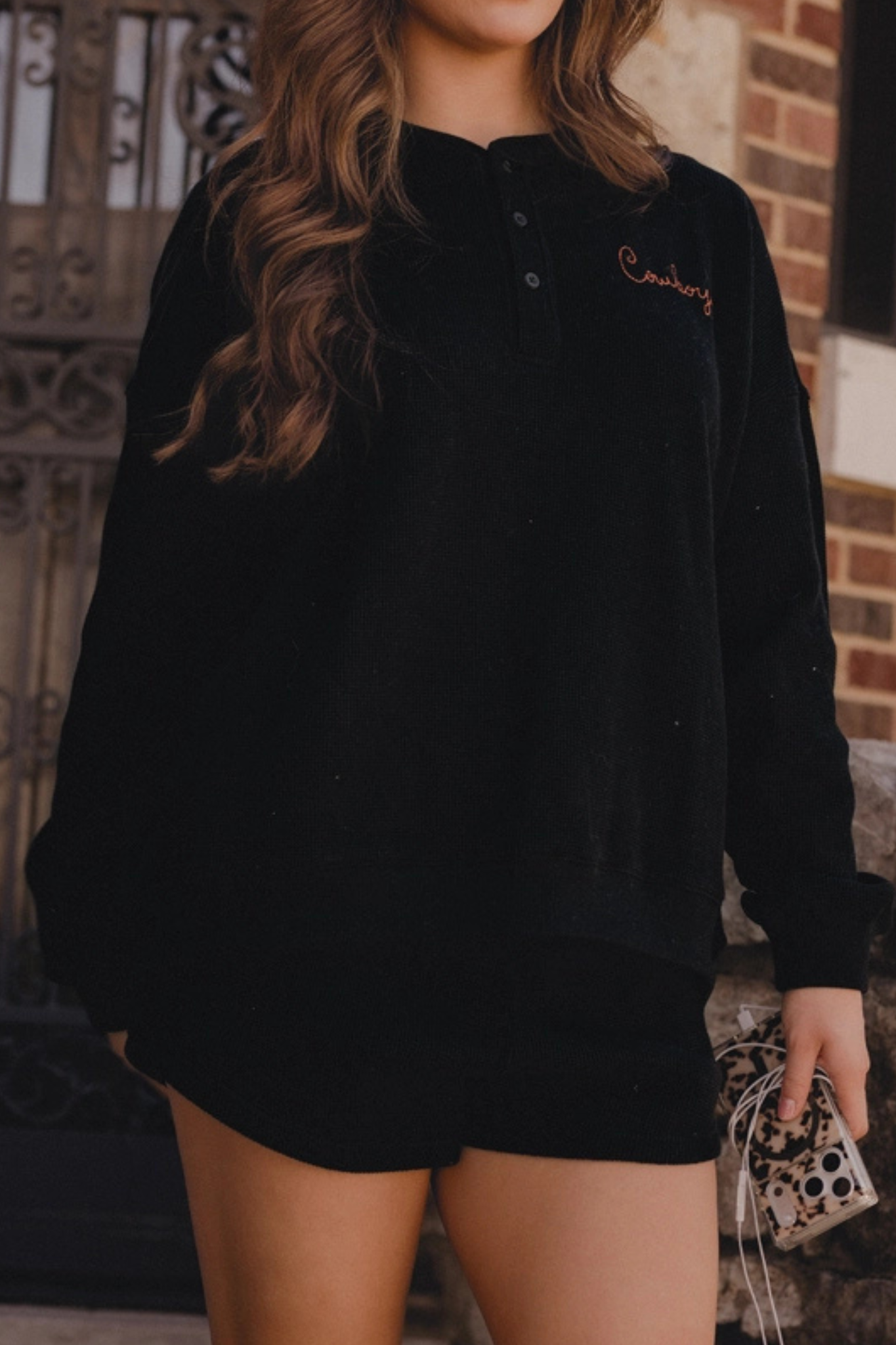 A woman wearing a black long-sleeve henley sweatshirt with a small embroidered script logo on the left chest, paired with black shorts, standing outdoors in front of a brick wall and metal gate.