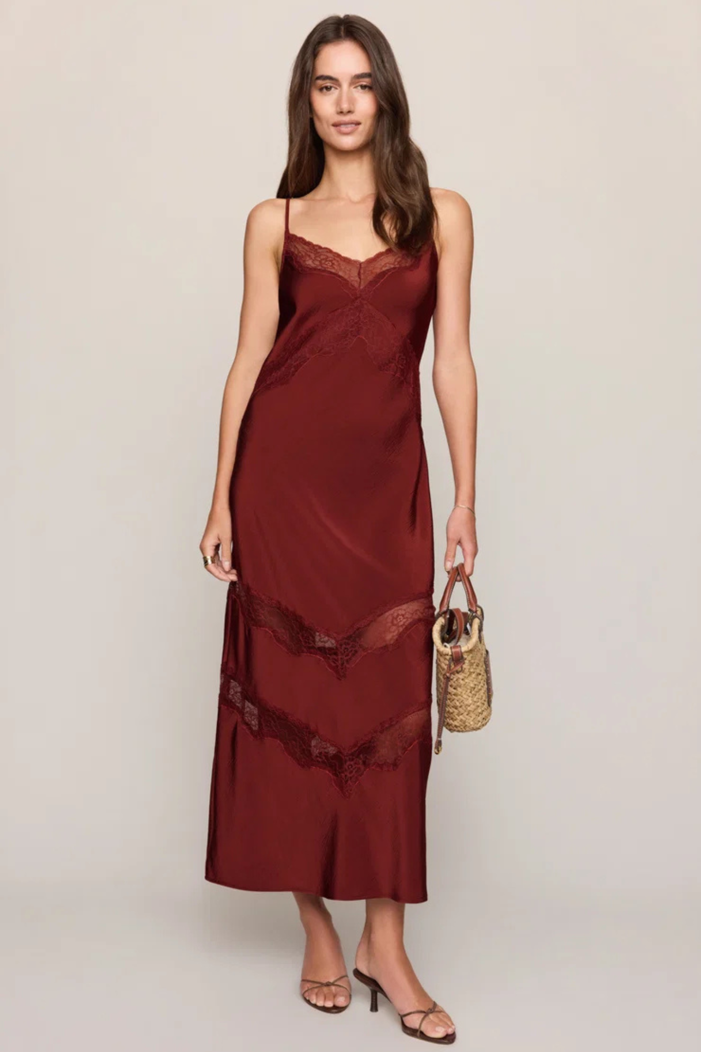 A woman modeling a long burgundy satin slip dress with lace trim, holding a small woven handbag against a plain light gray studio background.