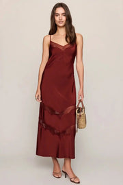 A woman modeling a long burgundy satin slip dress with lace trim, holding a small woven handbag against a plain light gray studio background.