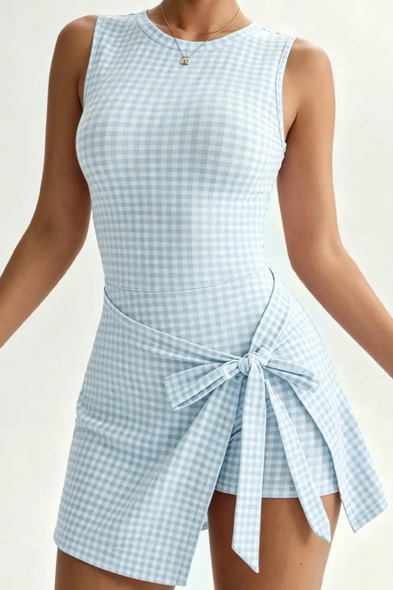 A sleeveless light blue and white gingham mini dress with an asymmetrical wrap skirt tied at the waist, shown on a model against a plain light background.