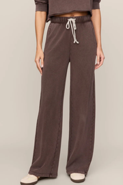 A person wearing wide-leg drawstring pants in a dark brown or taupe color, paired with a matching cropped top and light-colored shoes, against a plain beige studio background.